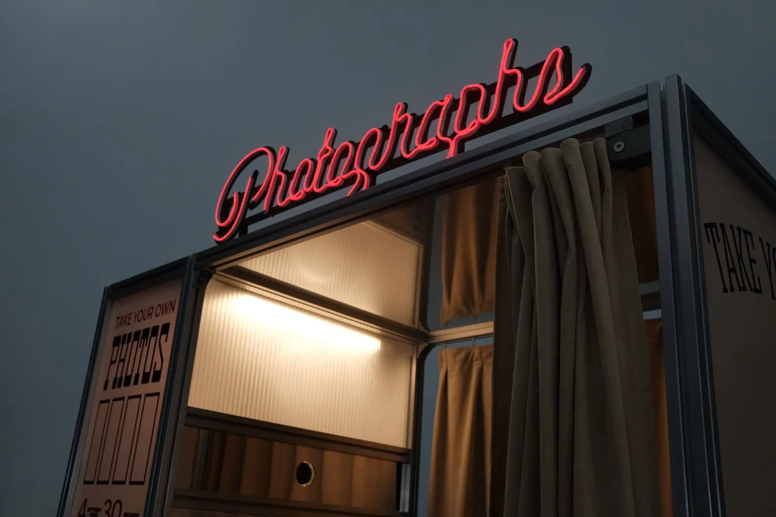 Close-up of the glowing red Photographs neon sign above the booth entrance at dusk